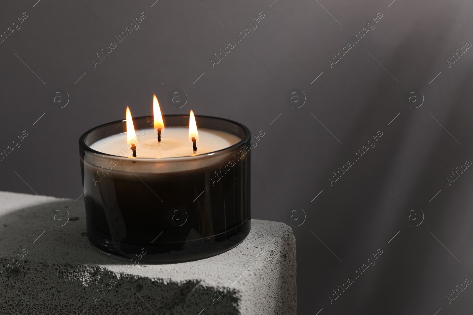 Photo of Burning soy wax candle on concrete block against gray background, space for text