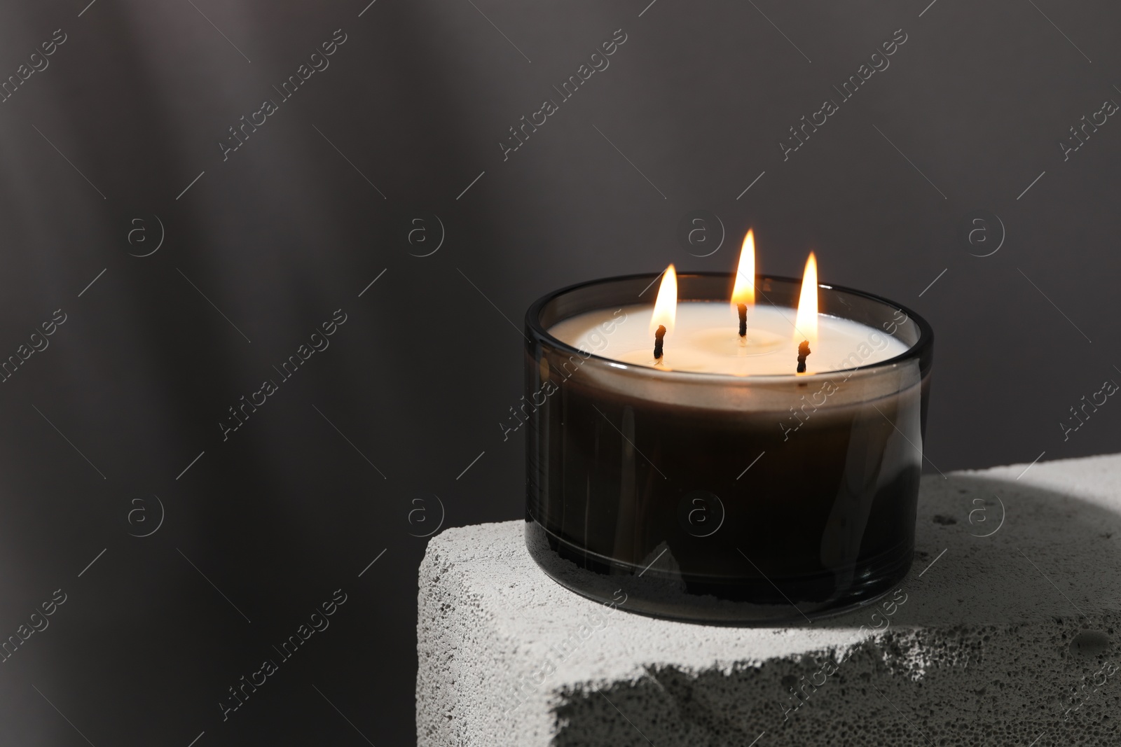 Photo of Burning soy wax candle on concrete block against gray background, space for text