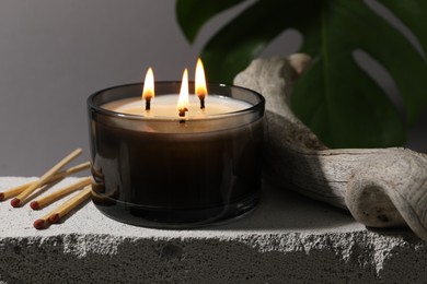 Photo of Burning soy wax candle, matches and decorative piece of wood on concrete block against gray background, closeup