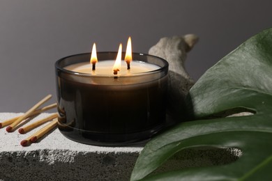 Photo of Burning soy wax candle, matches, decorative piece of wood and monstera leaf on concrete block against gray background, closeup