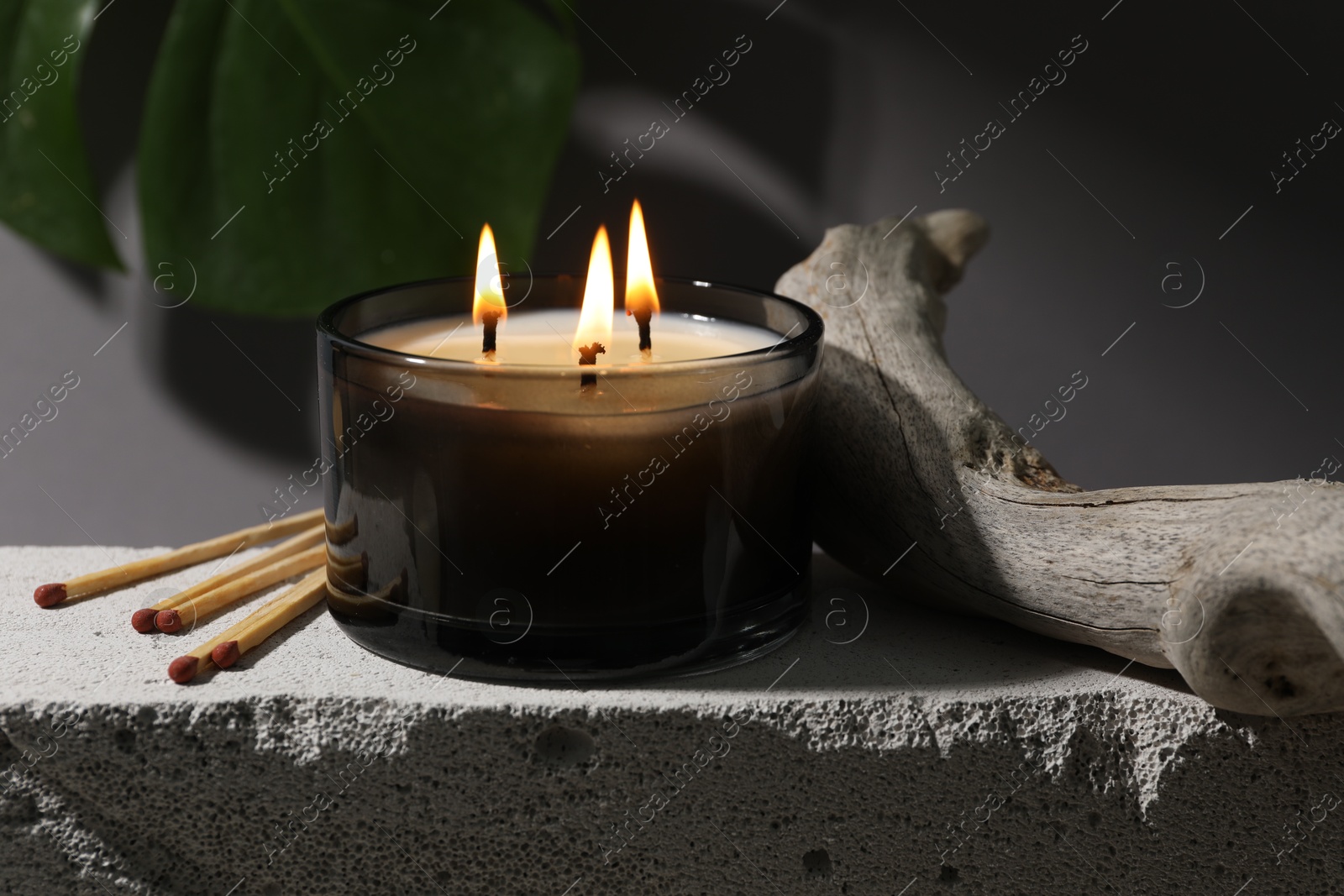 Photo of Burning soy wax candle, matches and decorative piece of wood on concrete block against gray background, closeup