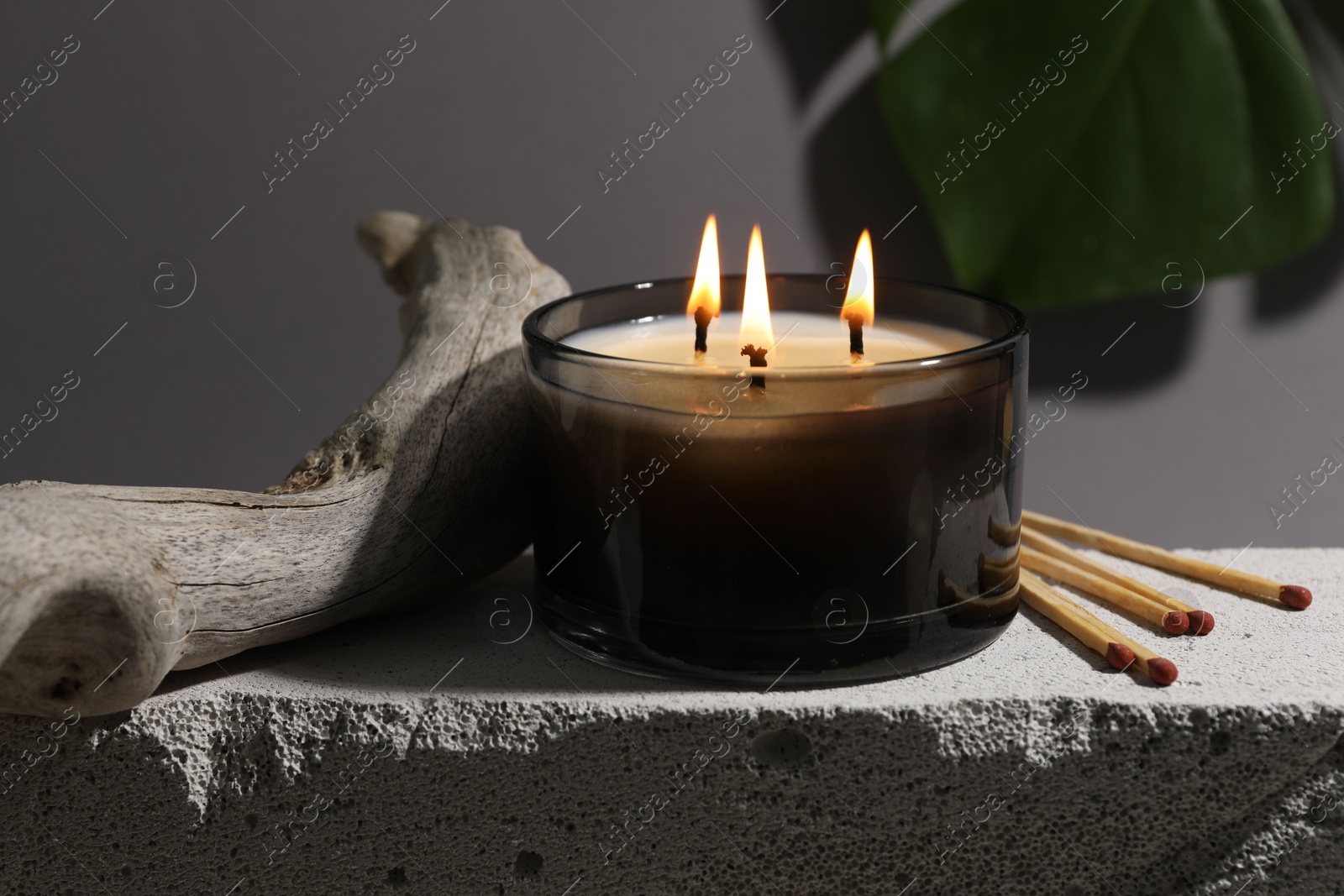 Photo of Burning soy wax candle, matches and decorative piece of wood on concrete block against gray background, closeup