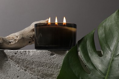 Photo of Burning soy wax candle on concrete block, decorative piece of wood and monstera leaf against gray background