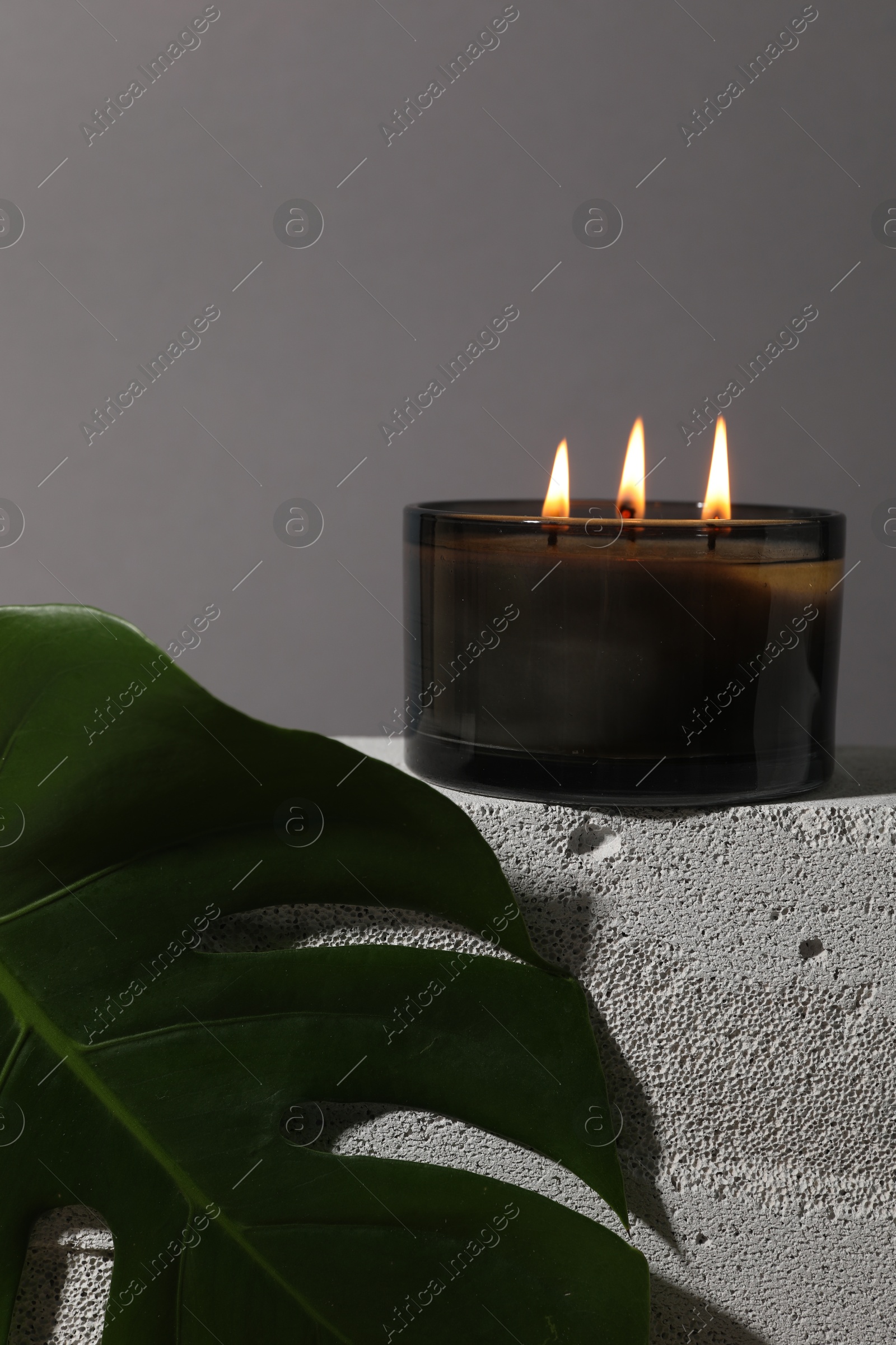 Photo of Burning soy wax candle on concrete block and monstera leaf against gray background, space for text