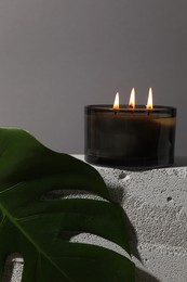 Photo of Burning soy wax candle on concrete block and monstera leaf against gray background, space for text