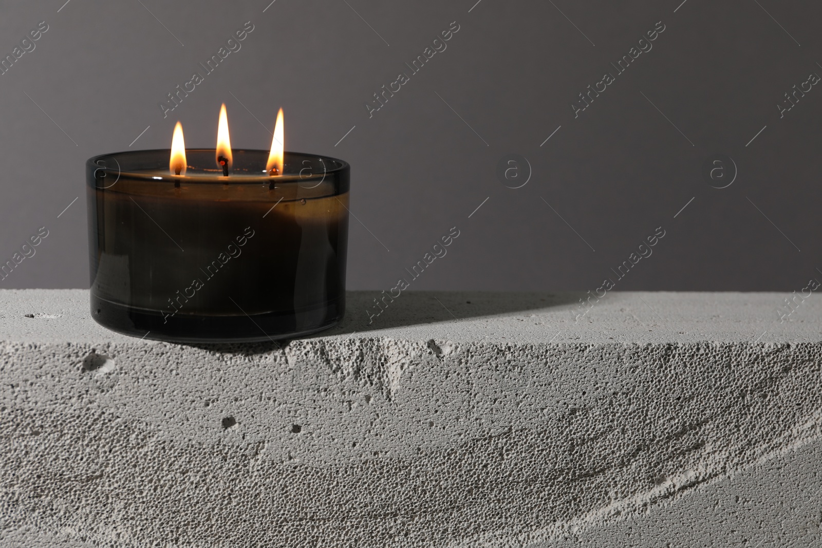 Photo of Burning soy wax candle on concrete block against gray background, space for text