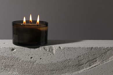 Photo of Burning soy wax candle on concrete block against gray background, space for text