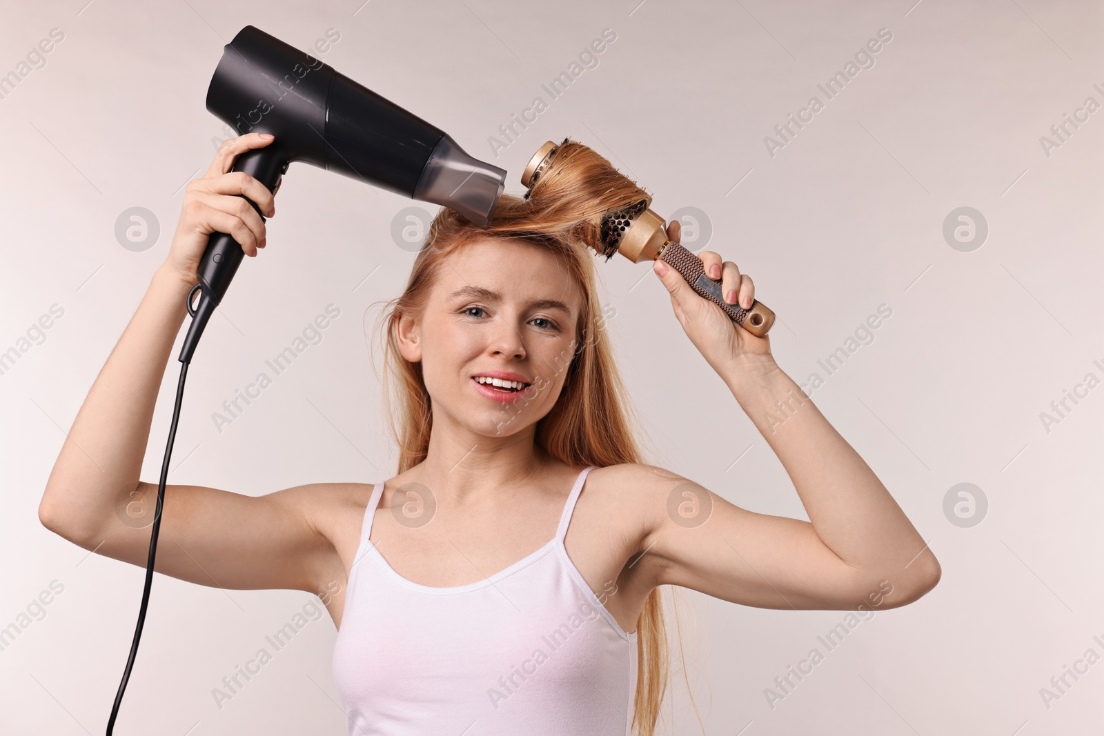 Photo of Beautiful young woman styling her hair with hairdryer and brush on light grey background