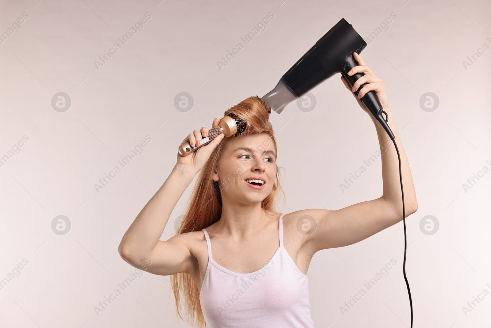 Photo of Beautiful young woman styling her hair with hairdryer and brush on light grey background