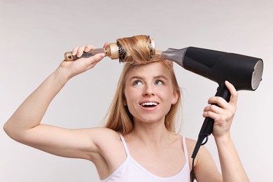 Photo of Beautiful young woman styling her hair with hairdryer and brush on light grey background