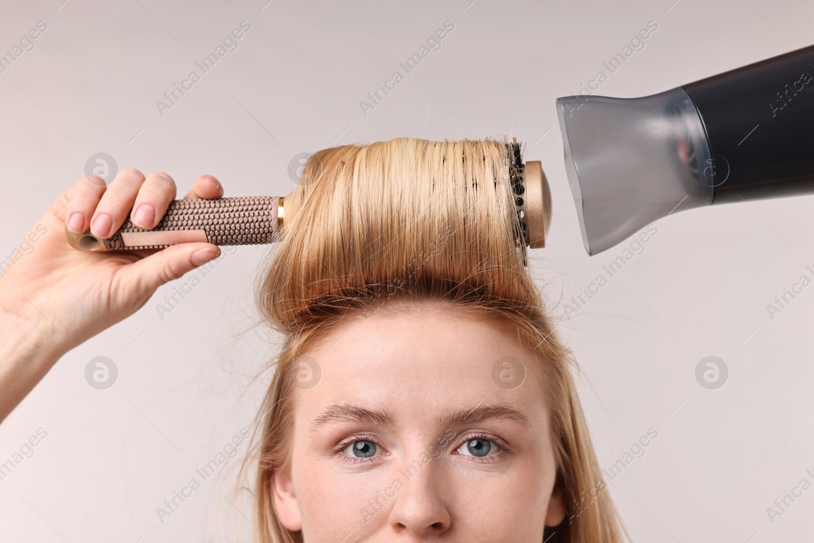 Photo of Beautiful young woman styling her hair with hairdryer and brush on light grey background, closeup