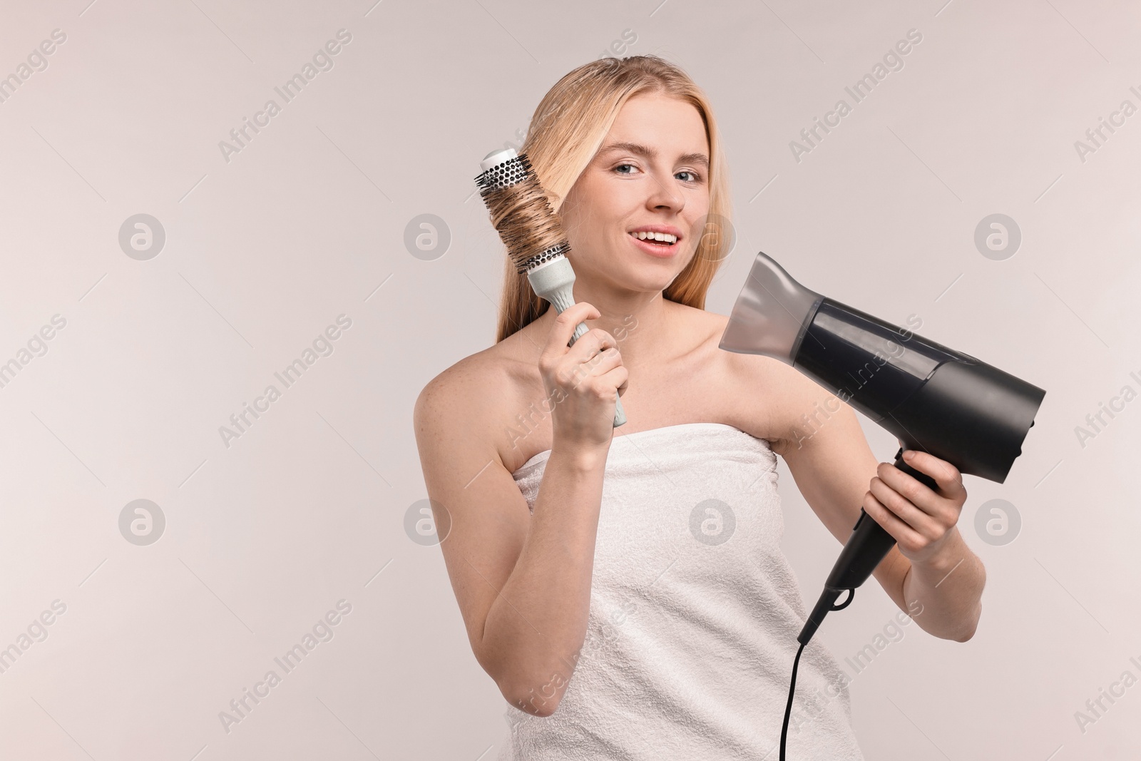 Photo of Beautiful young woman styling her hair with hairdryer and brush on light grey background