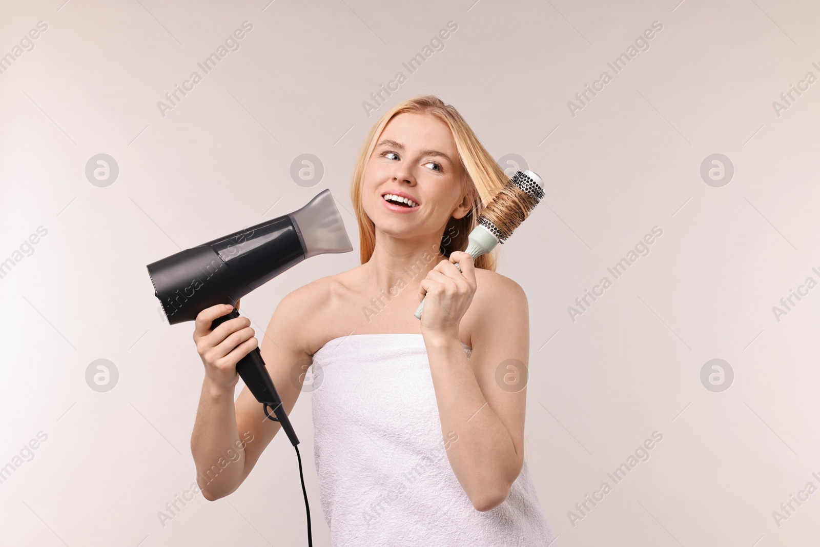 Photo of Beautiful young woman styling her hair with hairdryer and brush on light grey background