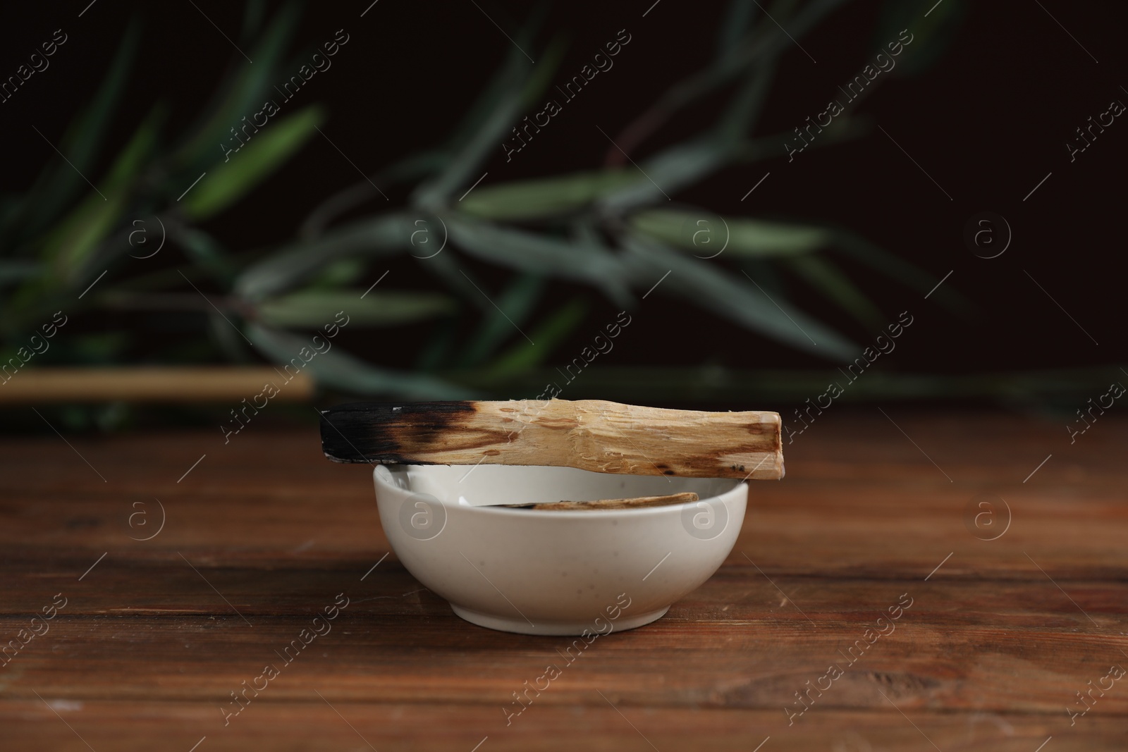 Photo of Burnt palo santo stick and bowl on wooden table against black background, closeup