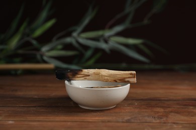 Photo of Burnt palo santo stick and bowl on wooden table against black background, closeup