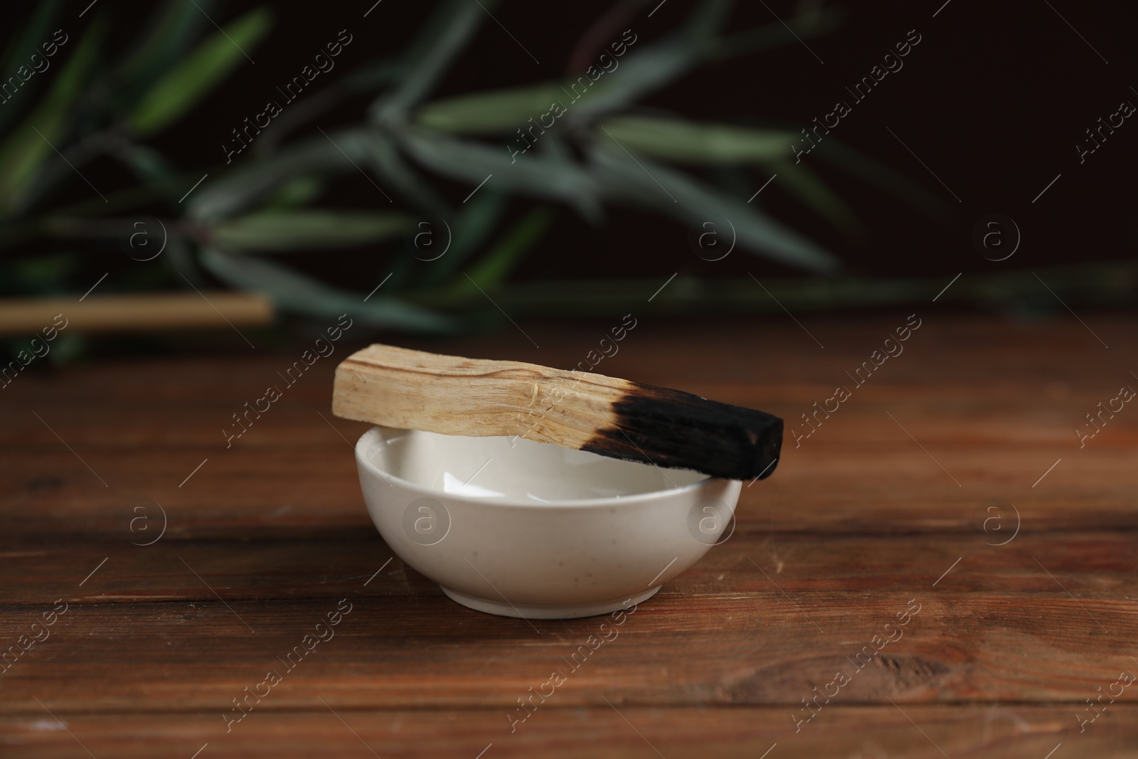 Photo of Burnt palo santo stick and bowl on wooden table against black background, closeup