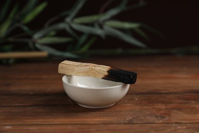 Photo of Burnt palo santo stick and bowl on wooden table against black background, closeup