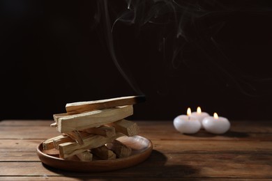 Photo of Palo santo sticks and smoldering one on wooden table against black background, closeup