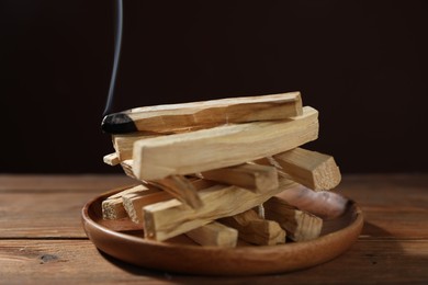 Photo of Palo santo sticks and smoldering one on wooden table against black background, closeup