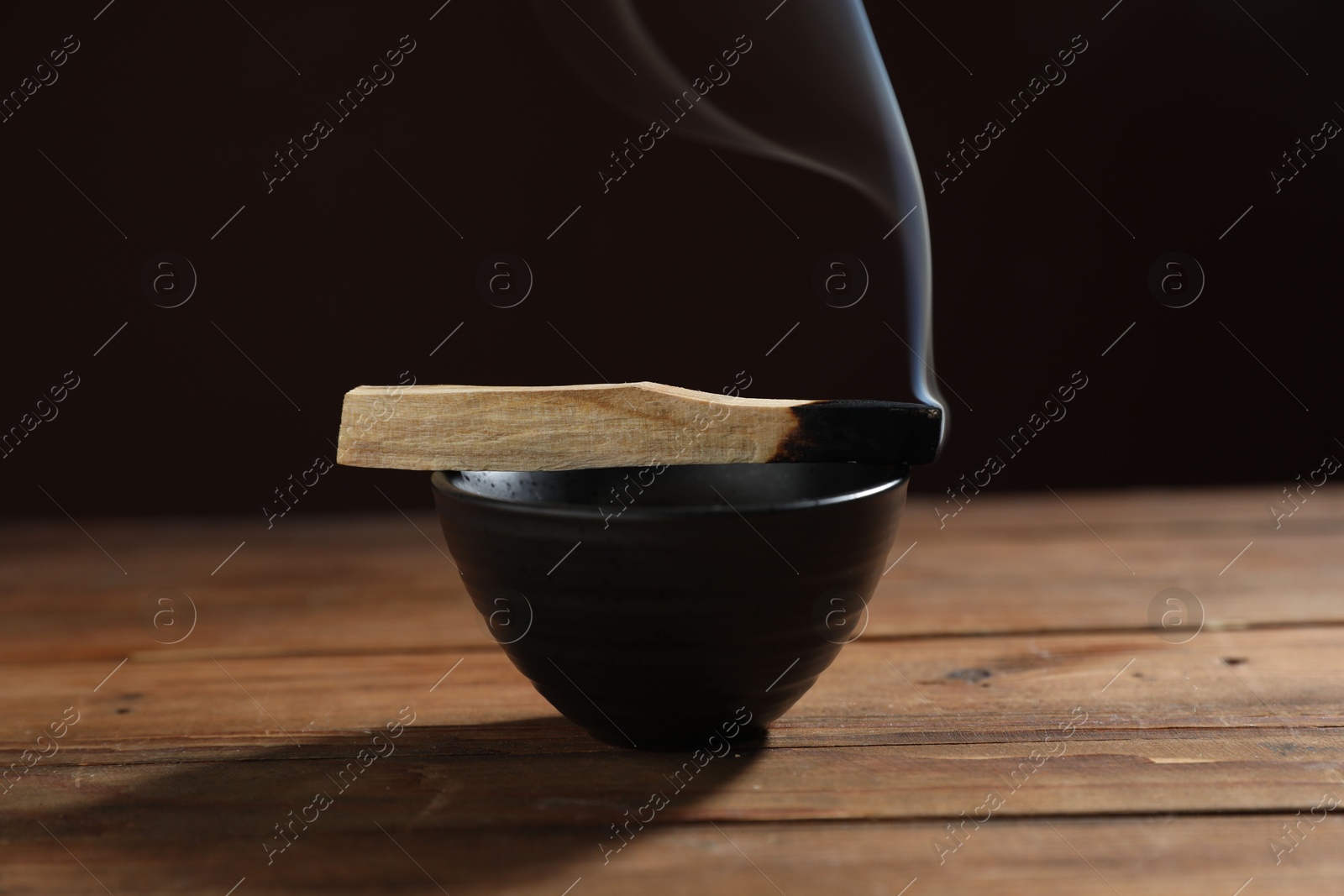 Photo of Smoldering palo santo stick and bowl on wooden table against black background, closeup