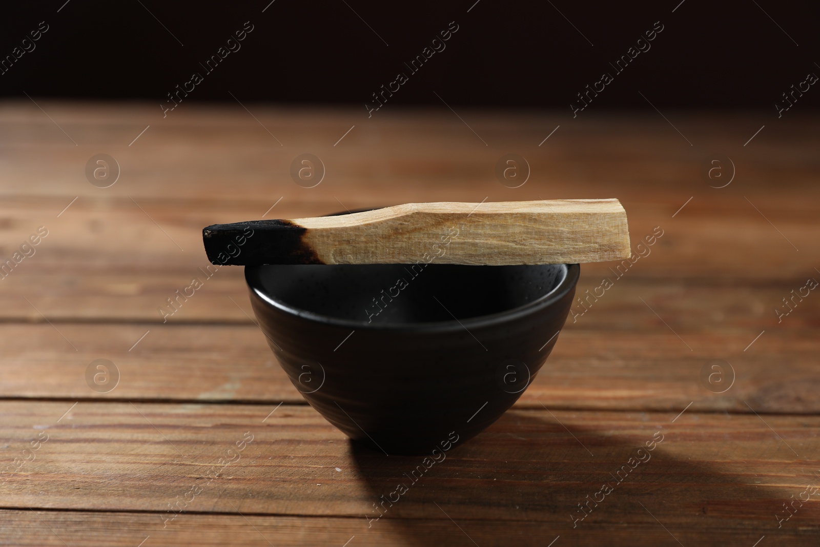 Photo of Burnt palo santo stick and bowl on wooden table against black background, closeup