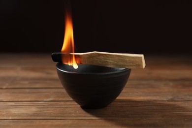 Photo of Burning palo santo stick and bowl on wooden table against black background, closeup