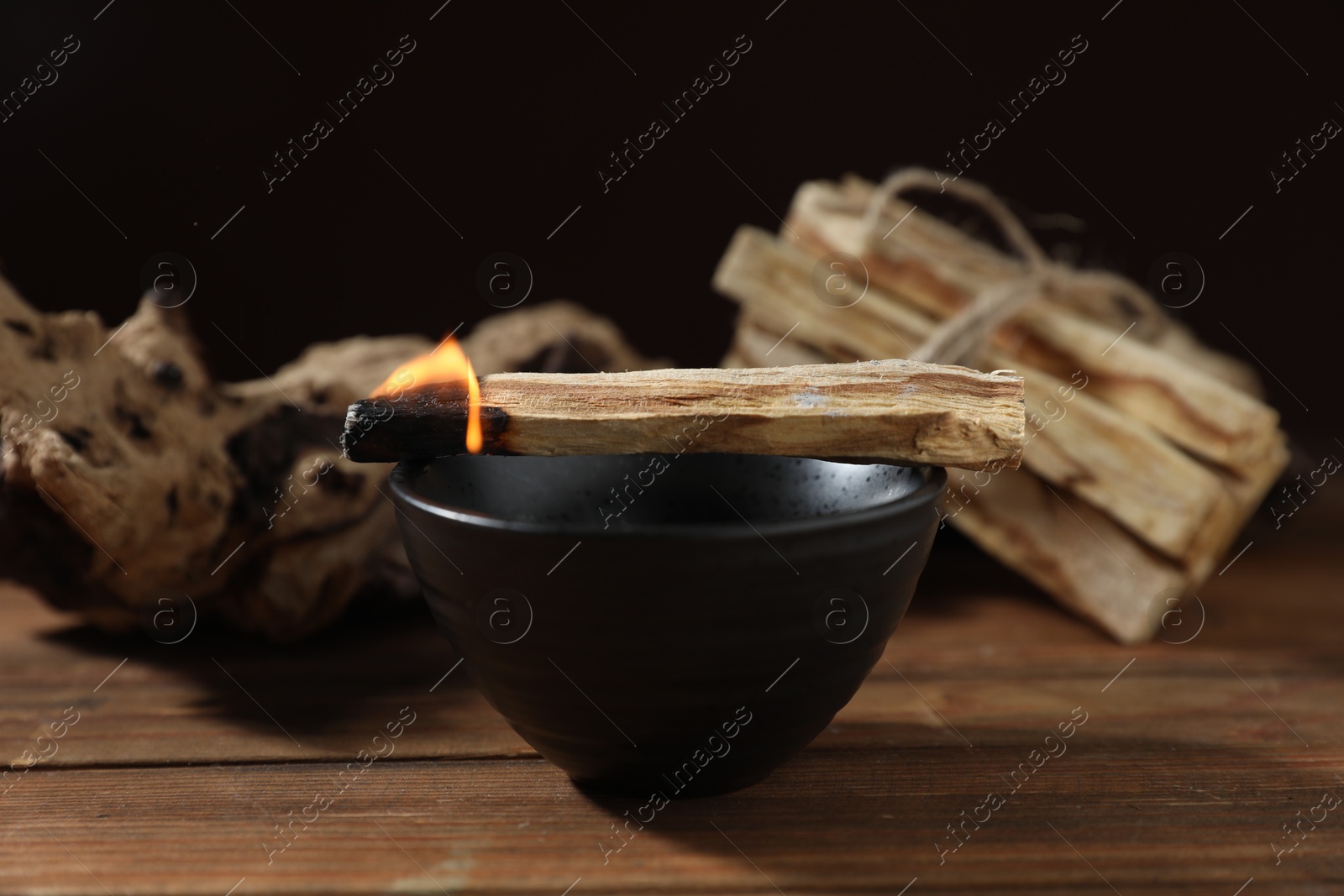 Photo of Burning palo santo stick and bowl on wooden table against black background, closeup