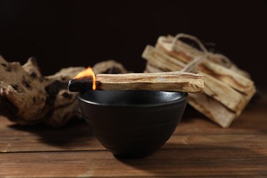 Photo of Burning palo santo stick and bowl on wooden table against black background, closeup