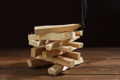 Photo of Palo santo sticks and smoldering one on wooden table against black background, closeup