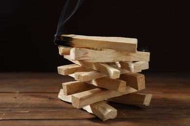 Photo of Palo santo sticks and smoldering one on wooden table against black background, closeup