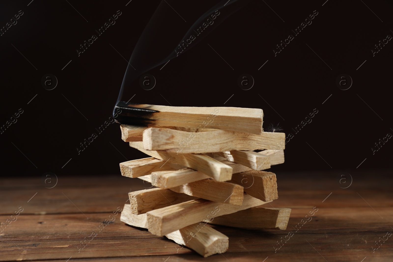 Photo of Palo santo sticks and smoldering one on wooden table against black background, closeup