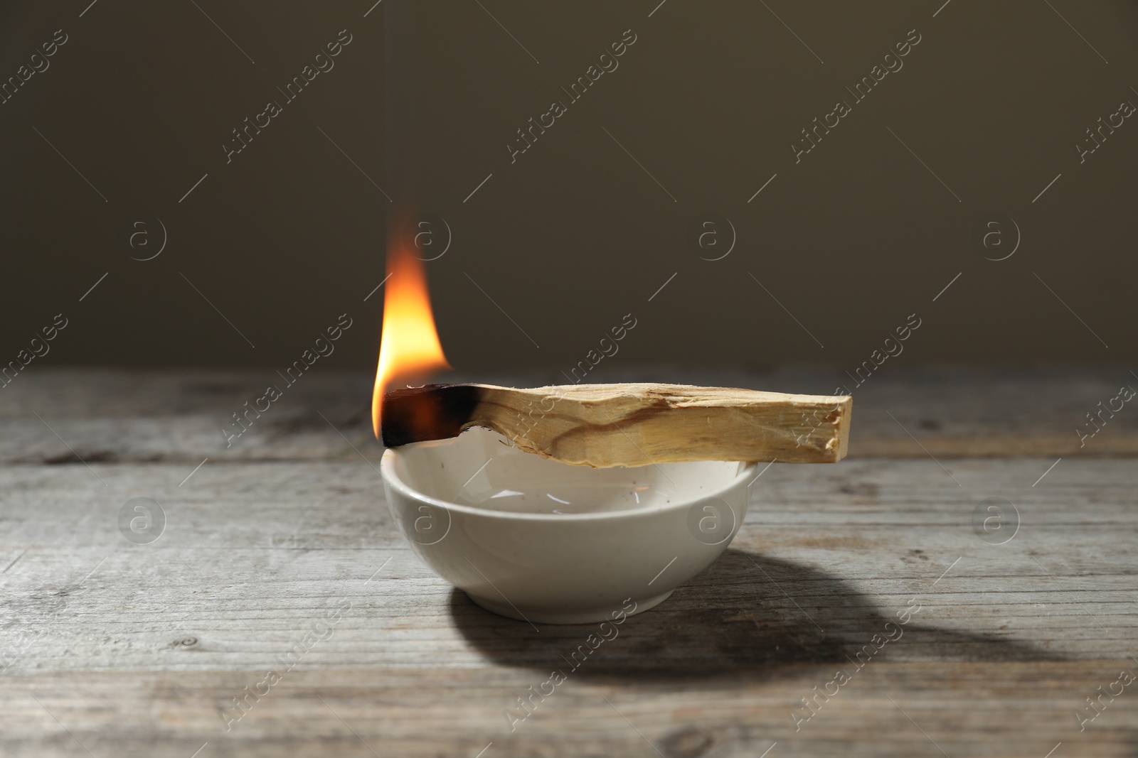 Photo of Burning palo santo stick and bowl on wooden table against grey background, closeup