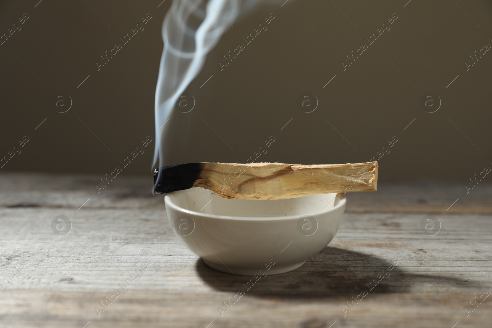 Photo of Smoldering palo santo stick and bowl on wooden table against grey background, closeup