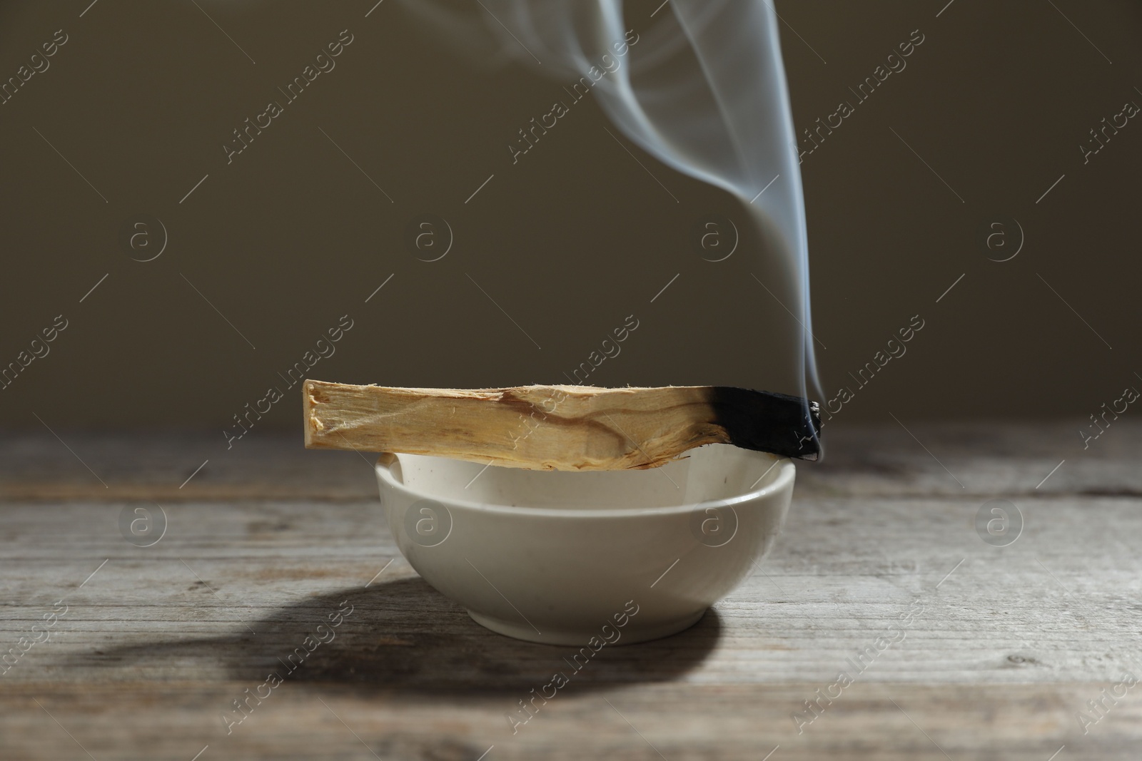 Photo of Smoldering palo santo stick and bowl on wooden table against grey background, closeup