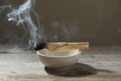 Photo of Smoldering palo santo stick and bowl on wooden table against grey background, closeup