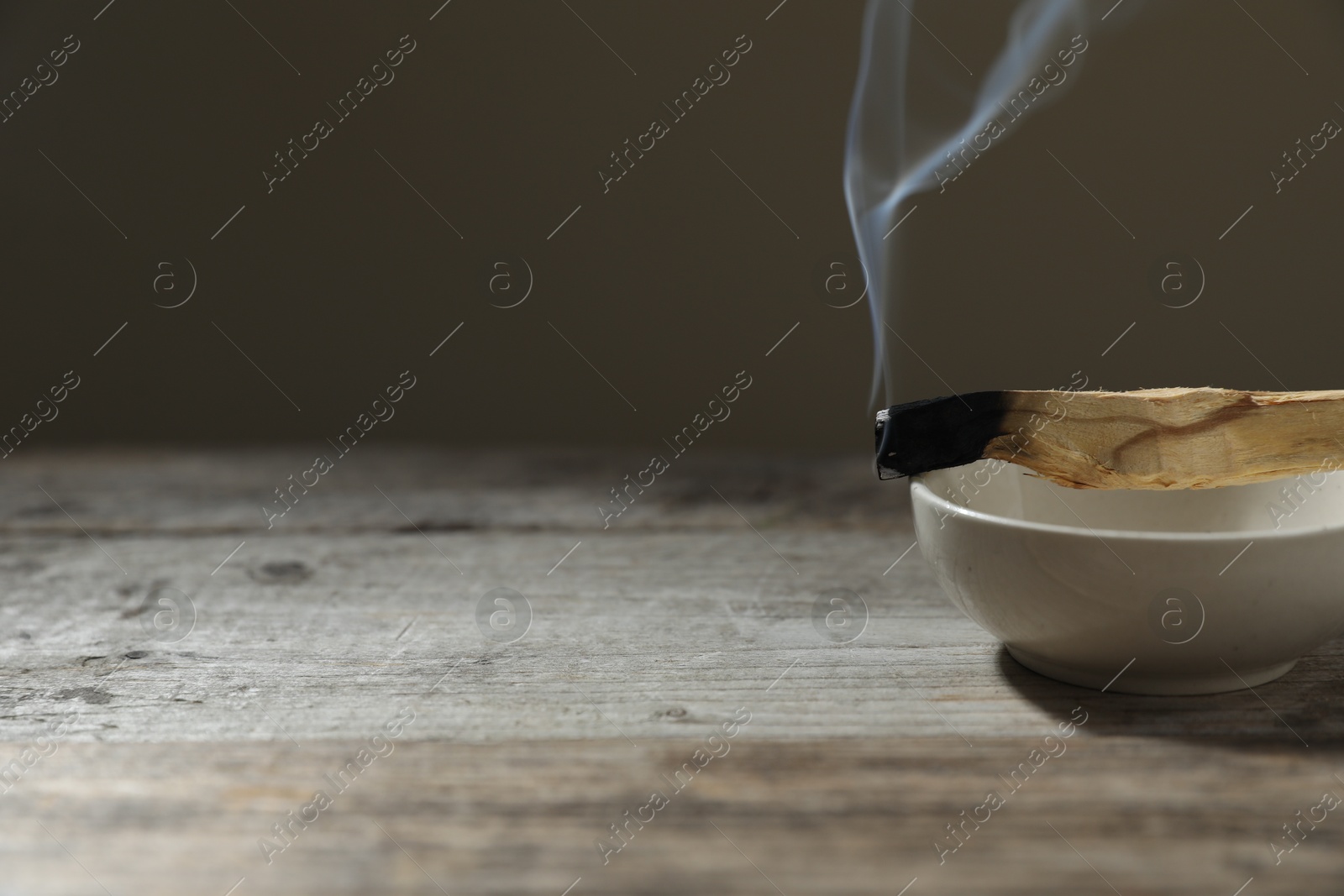 Photo of Smoldering palo santo stick and bowl on wooden table against grey background, closeup. Space for text