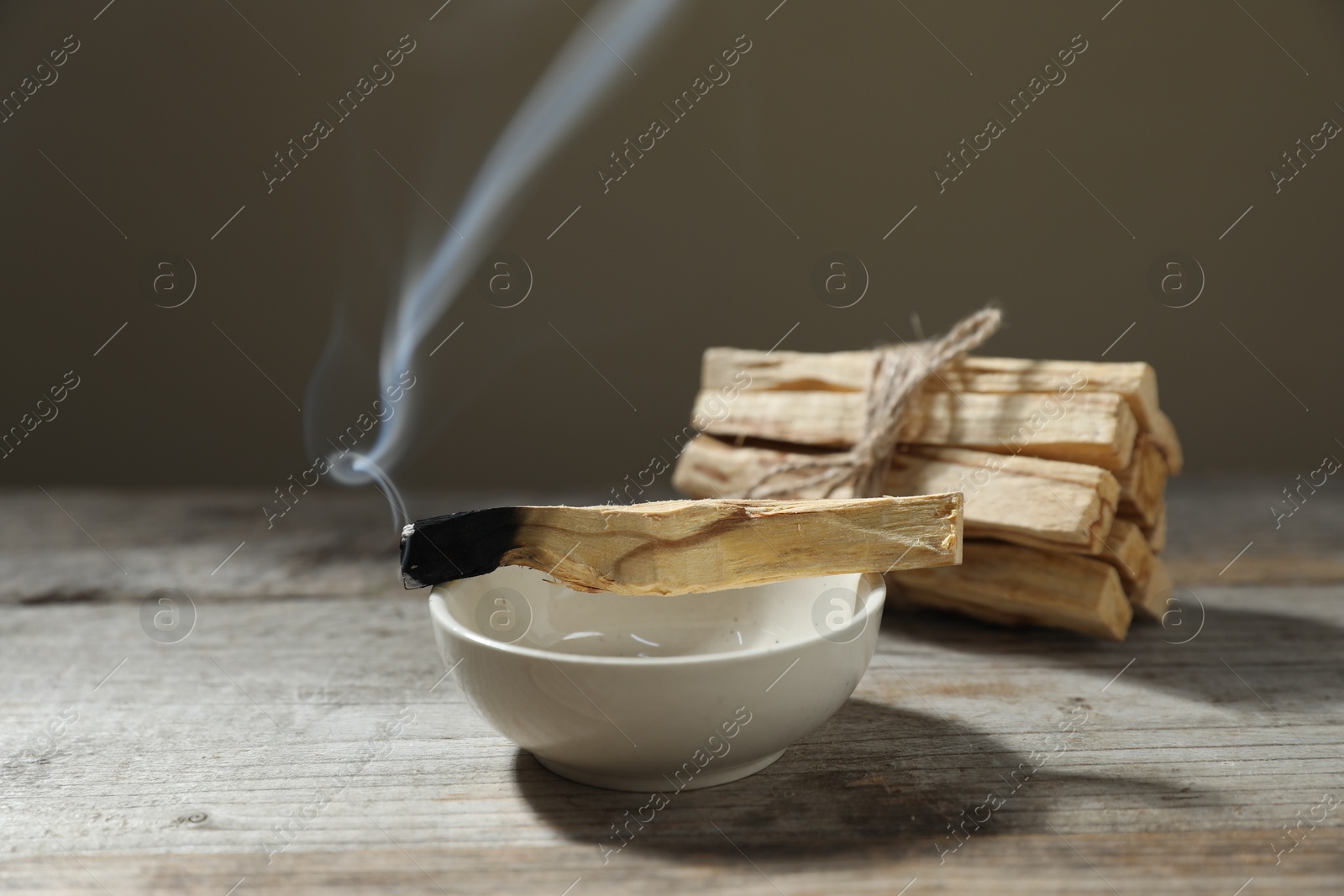 Photo of Smoldering palo santo stick and bowl on wooden table against grey background, closeup