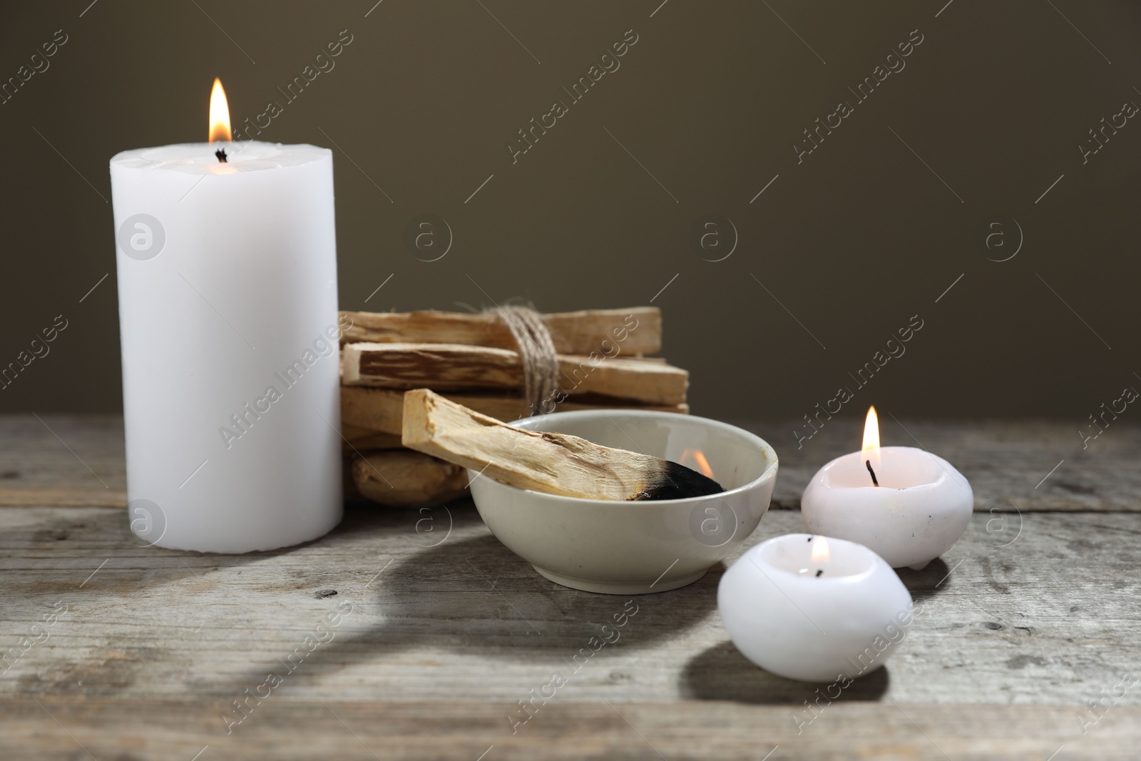 Photo of Burnt palo santo stick and burning candles on wooden table against grey background, closeup