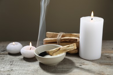 Photo of Smoldering palo santo stick and burning candles on wooden table against grey background, closeup