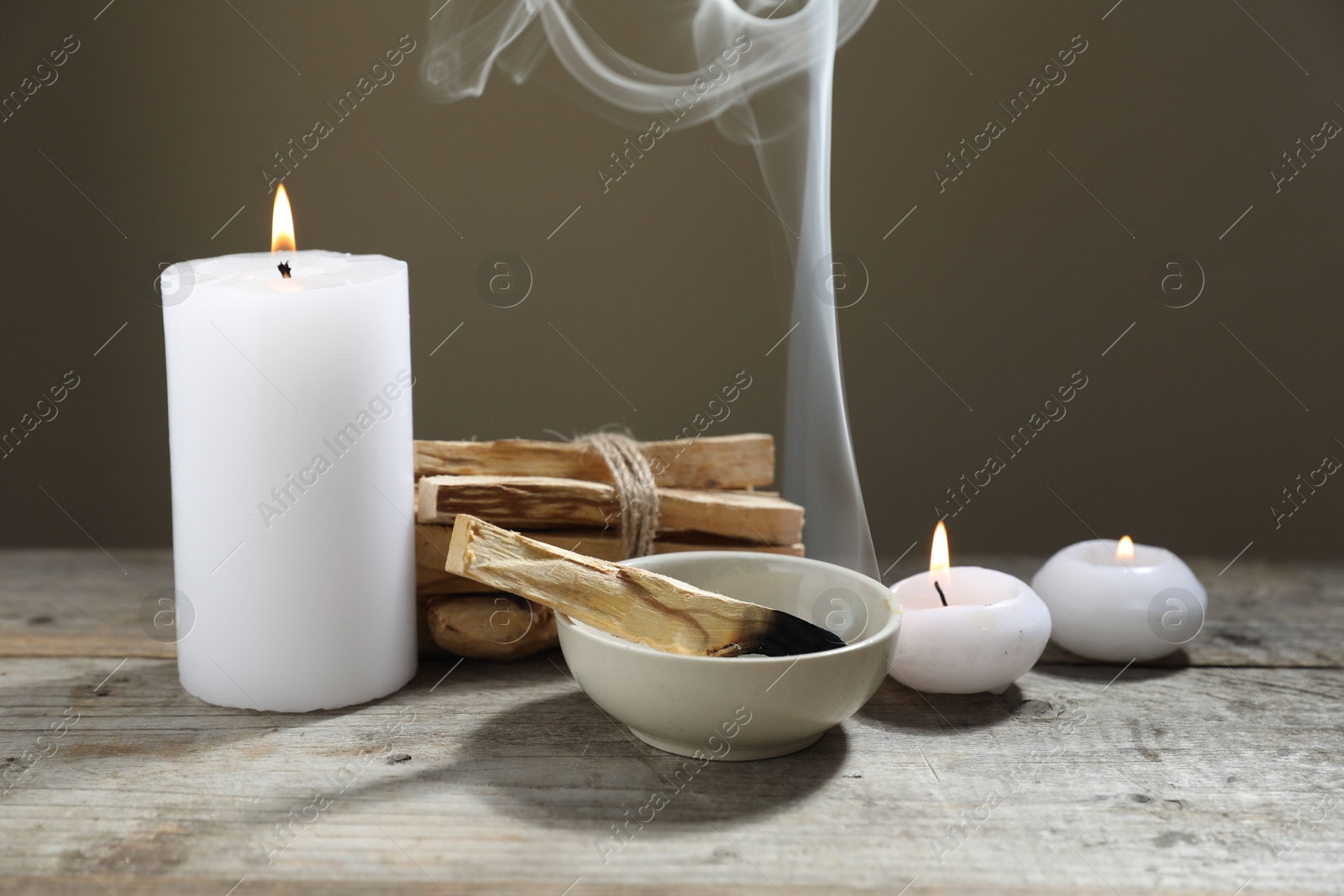 Photo of Smoldering palo santo stick and burning candles on wooden table against grey background, closeup