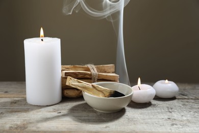 Photo of Smoldering palo santo stick and burning candles on wooden table against grey background, closeup