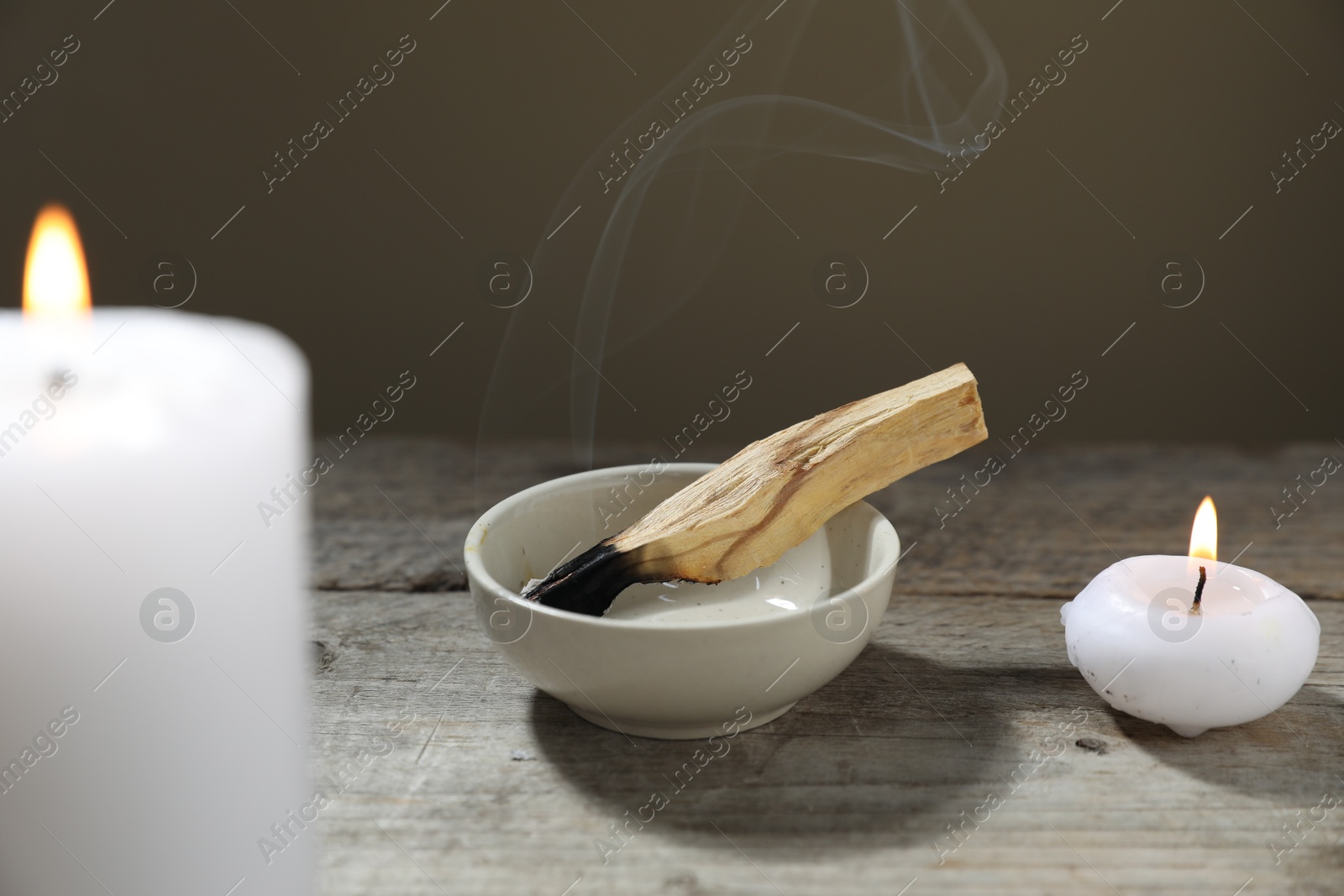 Photo of Smoldering palo santo stick and burning candles on wooden table against grey background, closeup