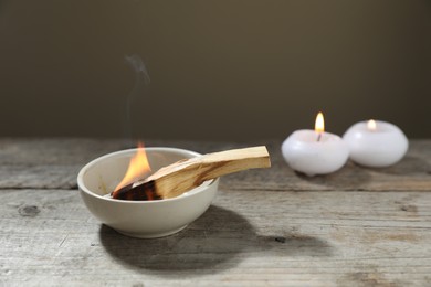 Photo of Burning palo santo stick and candles on wooden table against grey background, closeup