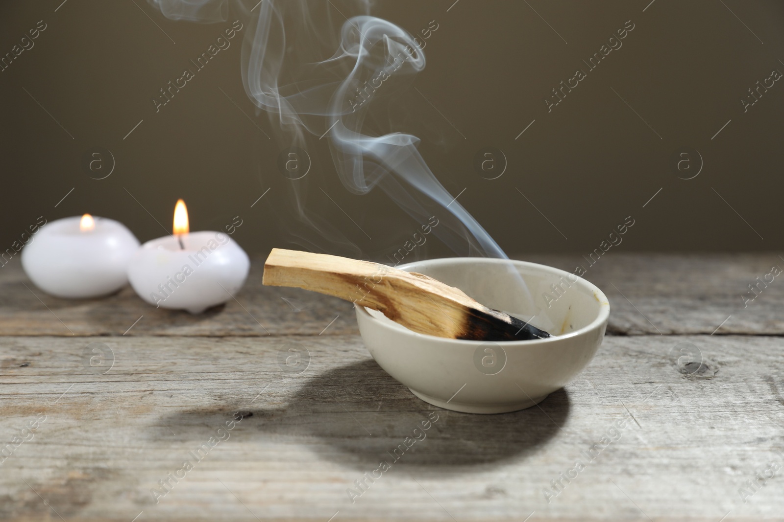 Photo of Smoldering palo santo stick and burning candles on wooden table against grey background, closeup