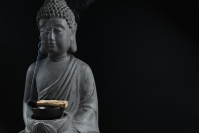 Photo of Smoldering palo santo stick and Buddha statue on black background, closeup. Space for text