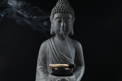 Photo of Smoldering palo santo stick and Buddha statue on black background, closeup