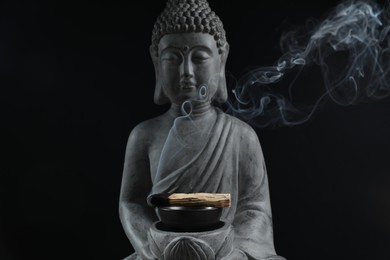 Photo of Smoldering palo santo stick and Buddha statue on black background, closeup