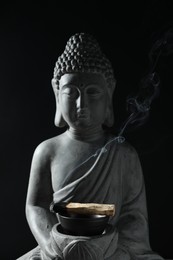 Photo of Smoldering palo santo stick and Buddha statue on black background, closeup