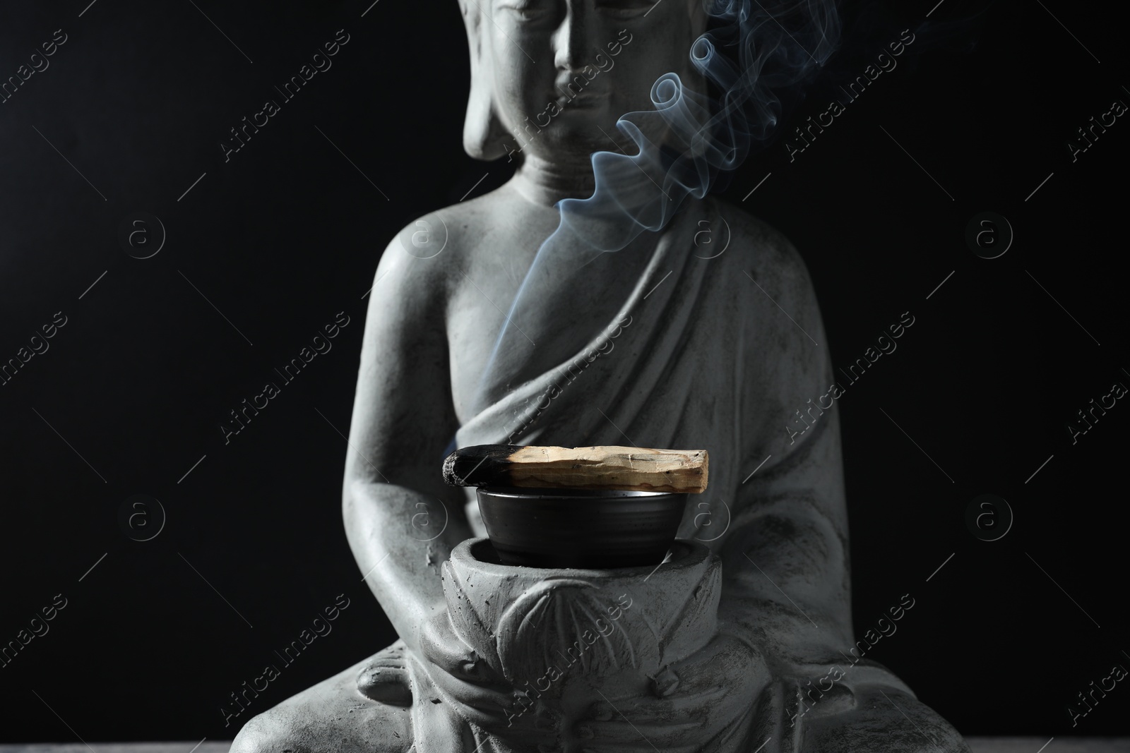 Photo of Smoldering palo santo stick and Buddha statue on black background, closeup