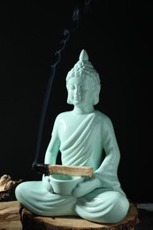 Photo of Smoldering palo santo stick and Buddha statue on table against black background, closeup